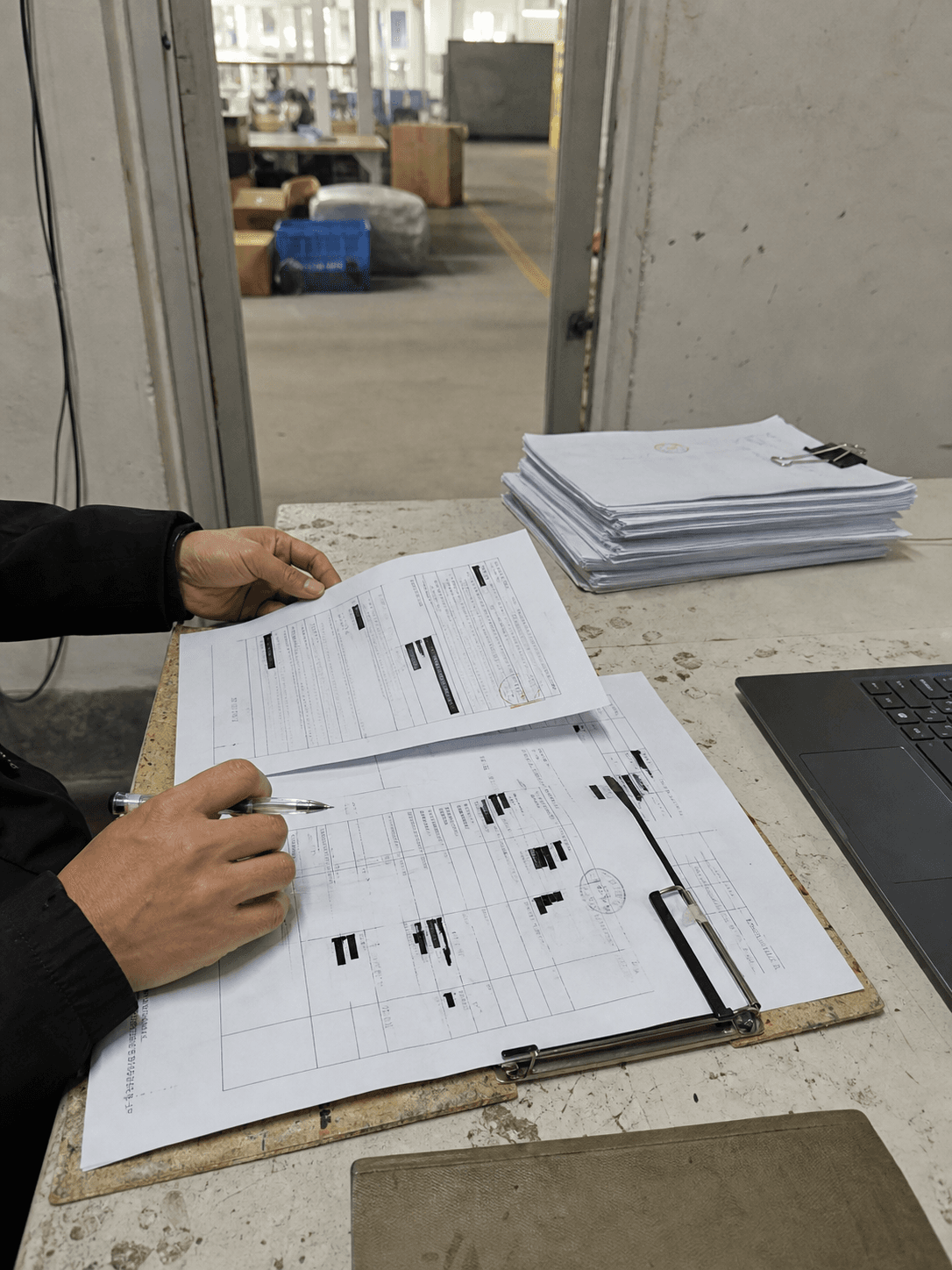 Hands reviewing redacted supplier audit paperwork on a worn factory office desk