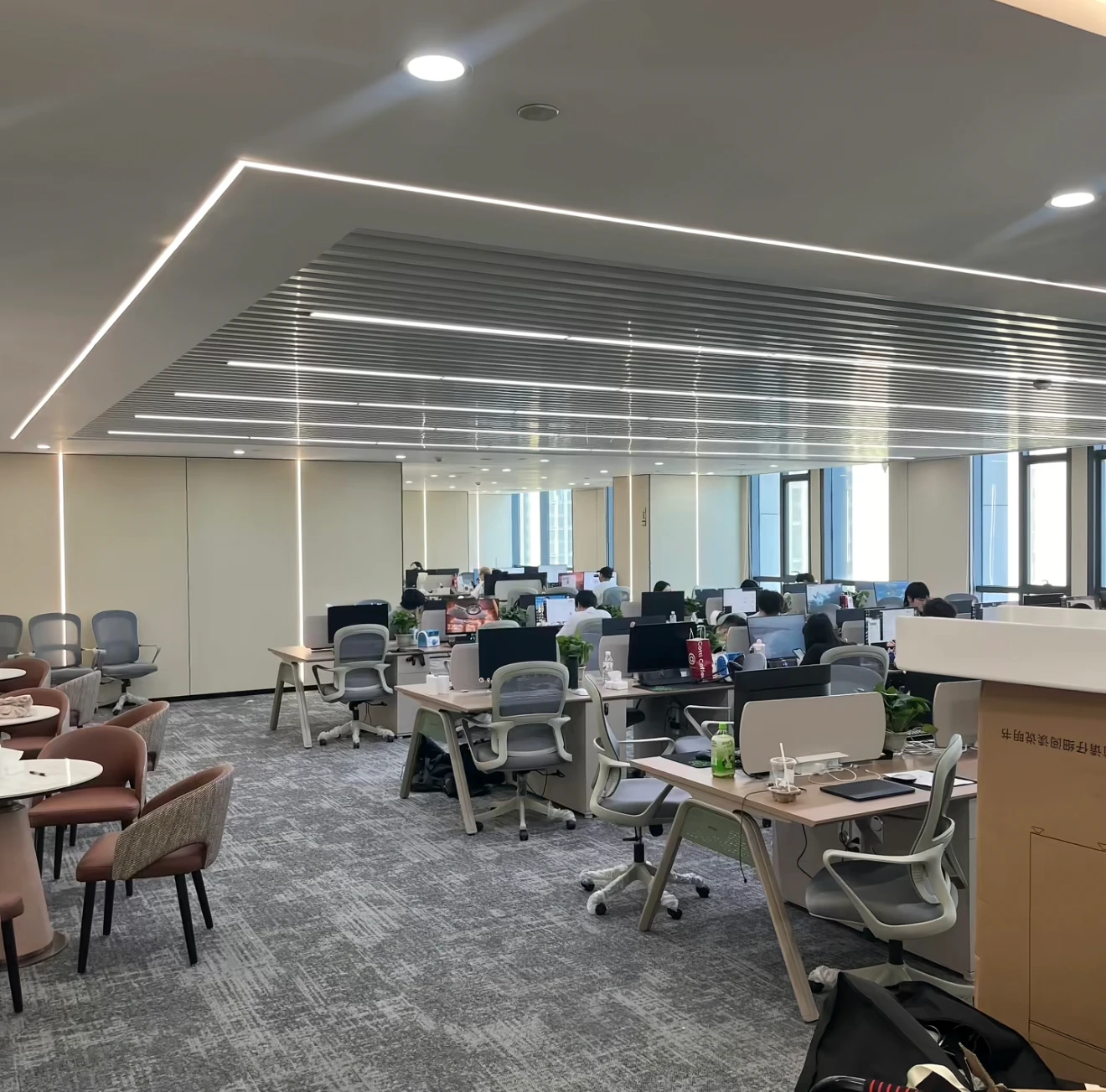Open-plan office with rows of desks, monitors and office chairs; on the left, a meeting area with round tables and chairs.