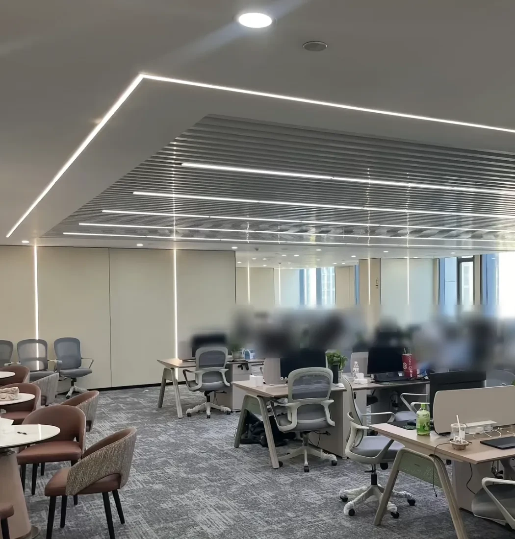 Open-plan office with rows of desks, monitors and office chairs; on the left, a meeting area with round tables and chairs.
